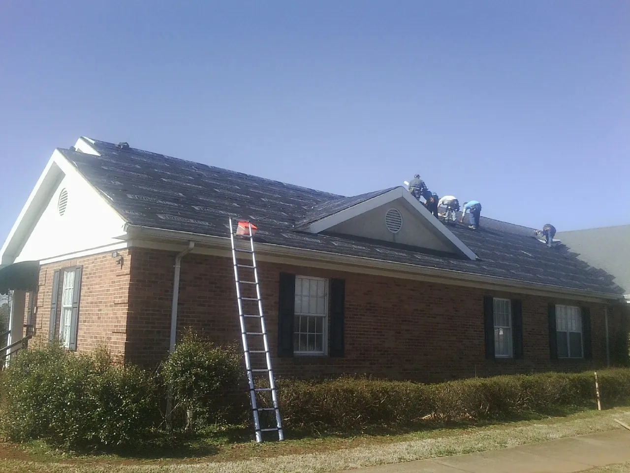 Crew tearing shingles off a brick ranch roof with a ladder set — Emergency Roof Repair work by our crew in Pine Hall