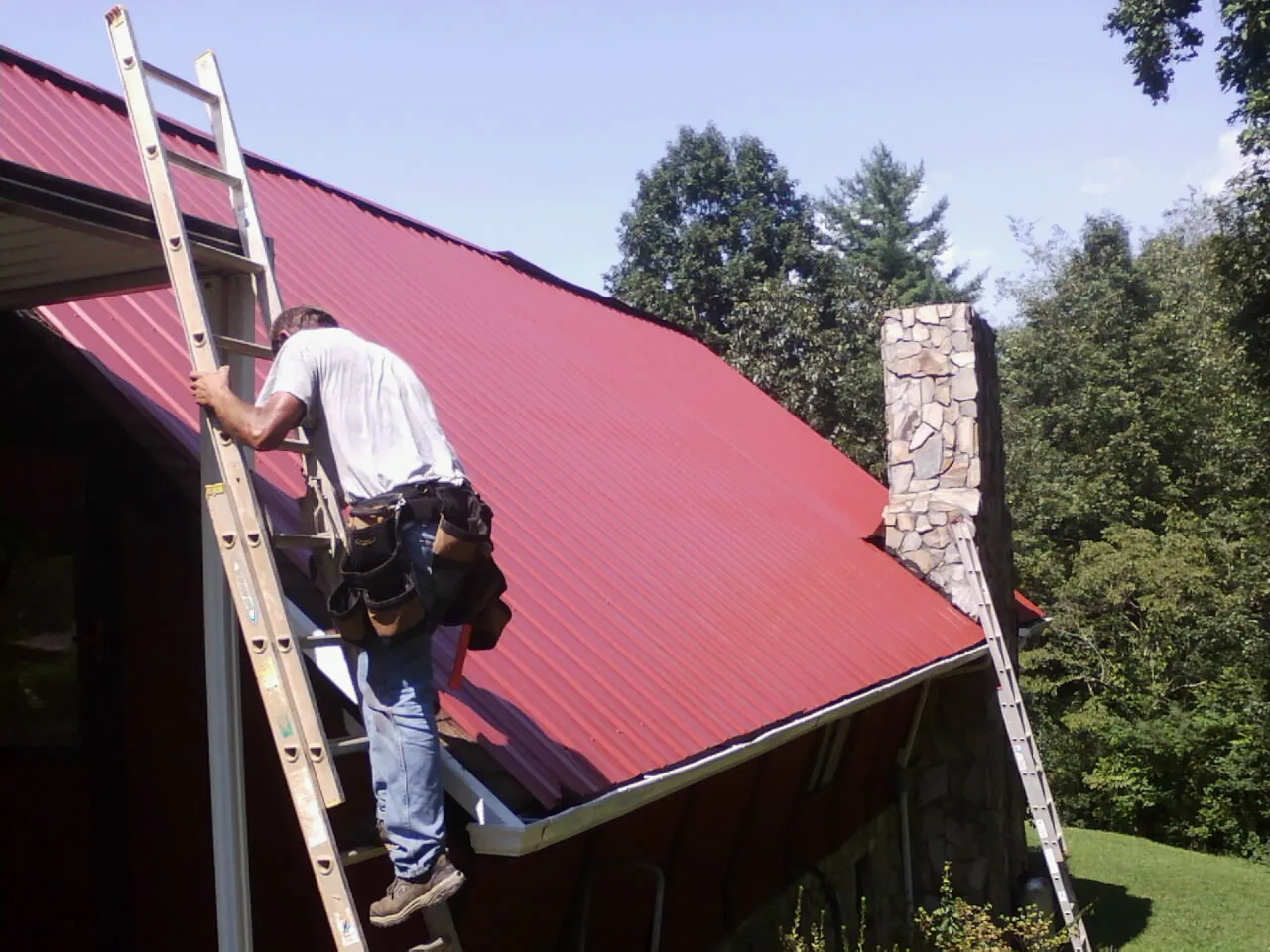 Roofer on a ladder beside a red metal roof and stone chimney — Soffit & Fascia Repair by Roofing Brothers in Pine Hall